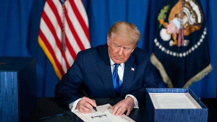 Donald Trump está sentado em frente a uma mesa, com uma caixa azul na lateral. Ele veste ternos escuros e assina um papel.